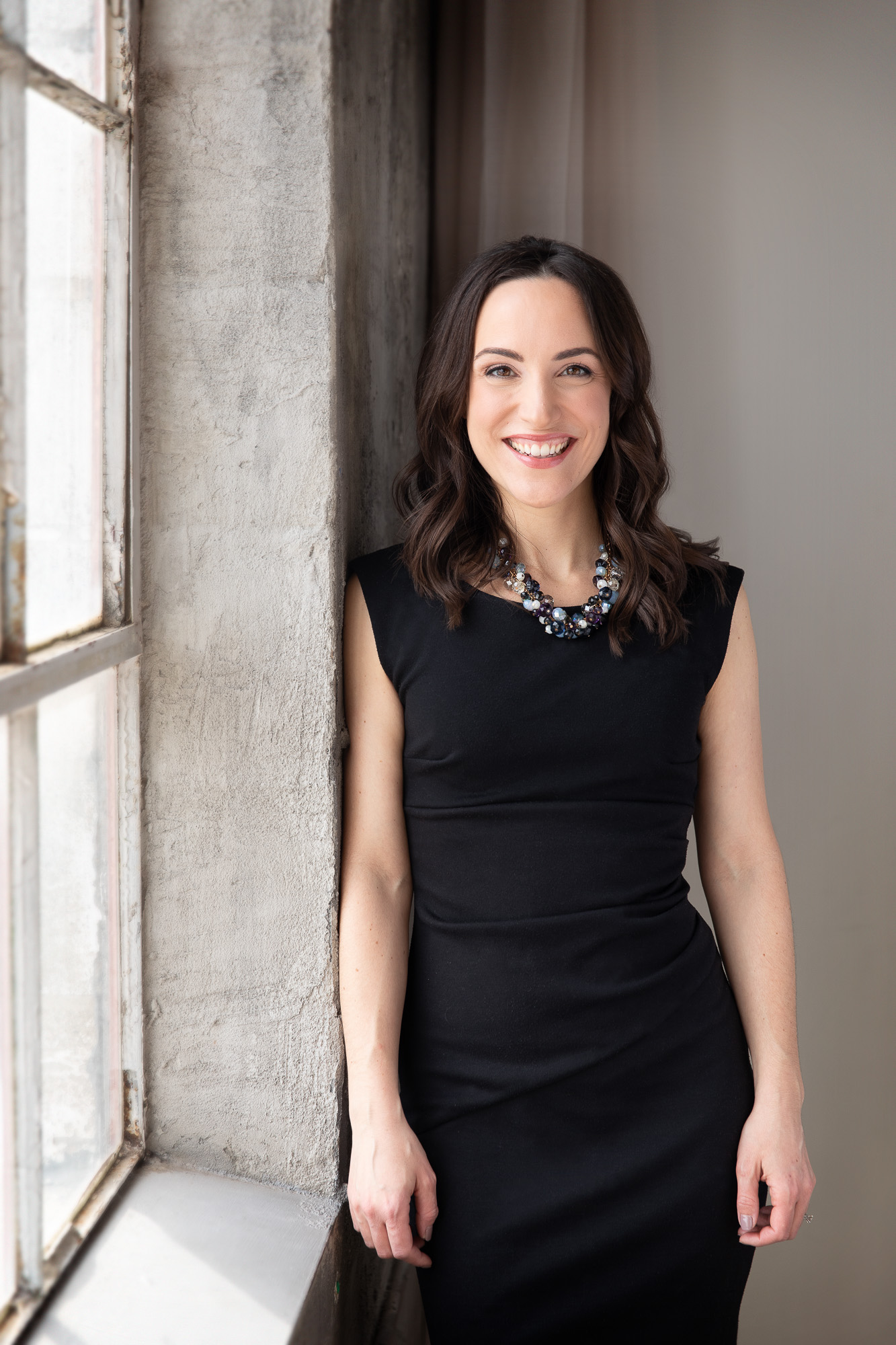 attractive woman with long dark hair and a black dress leaning agains the wall lit beautifully by the window light