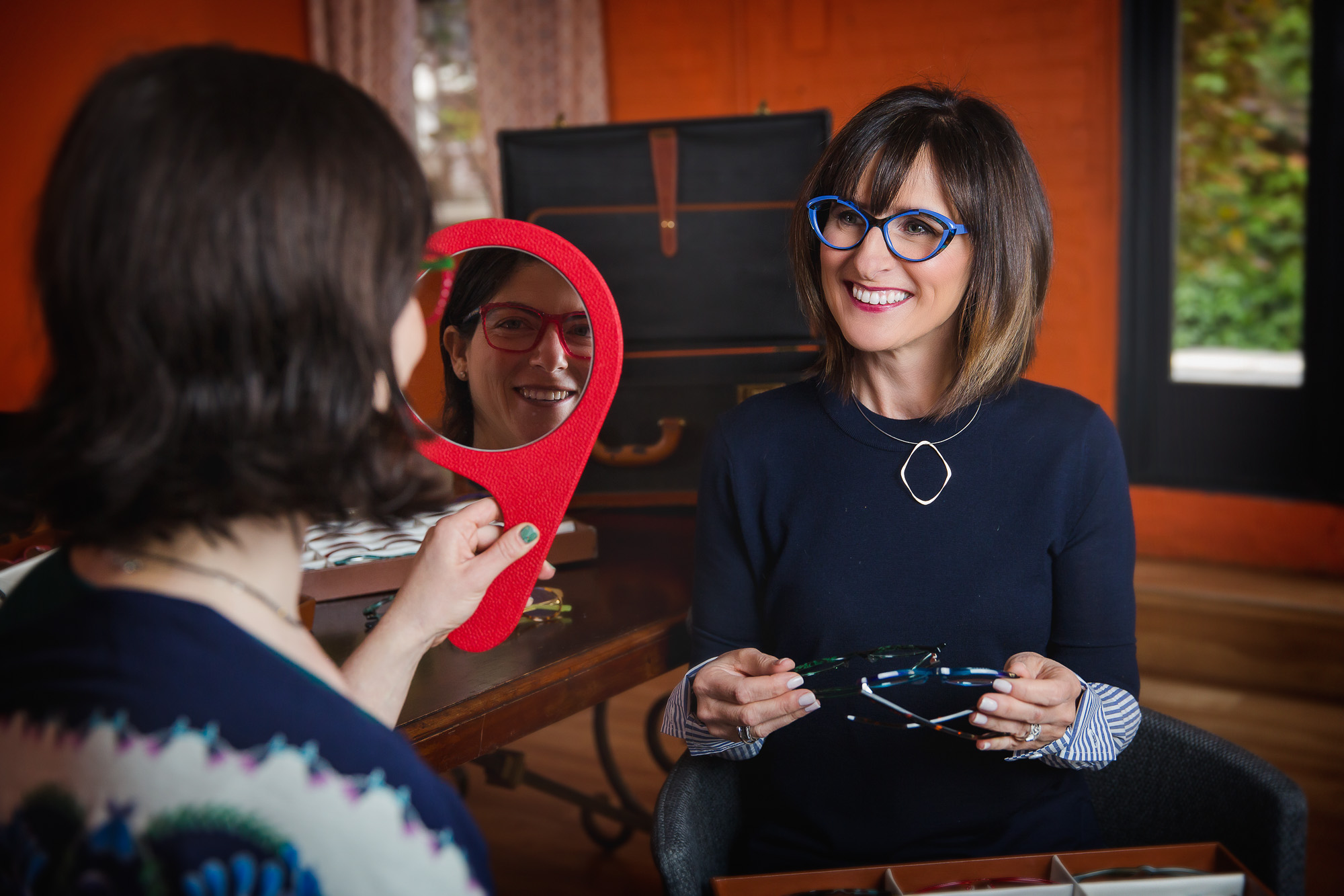 woman selling glasses to another woman who is trying them on and looking in the mirror